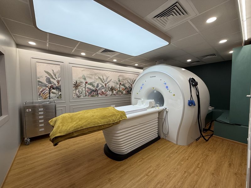 Wide-bore MRI scanner and its padded table in a softly lit room with a botanical wall mural at AV Scan Diagnostics, Dadar West